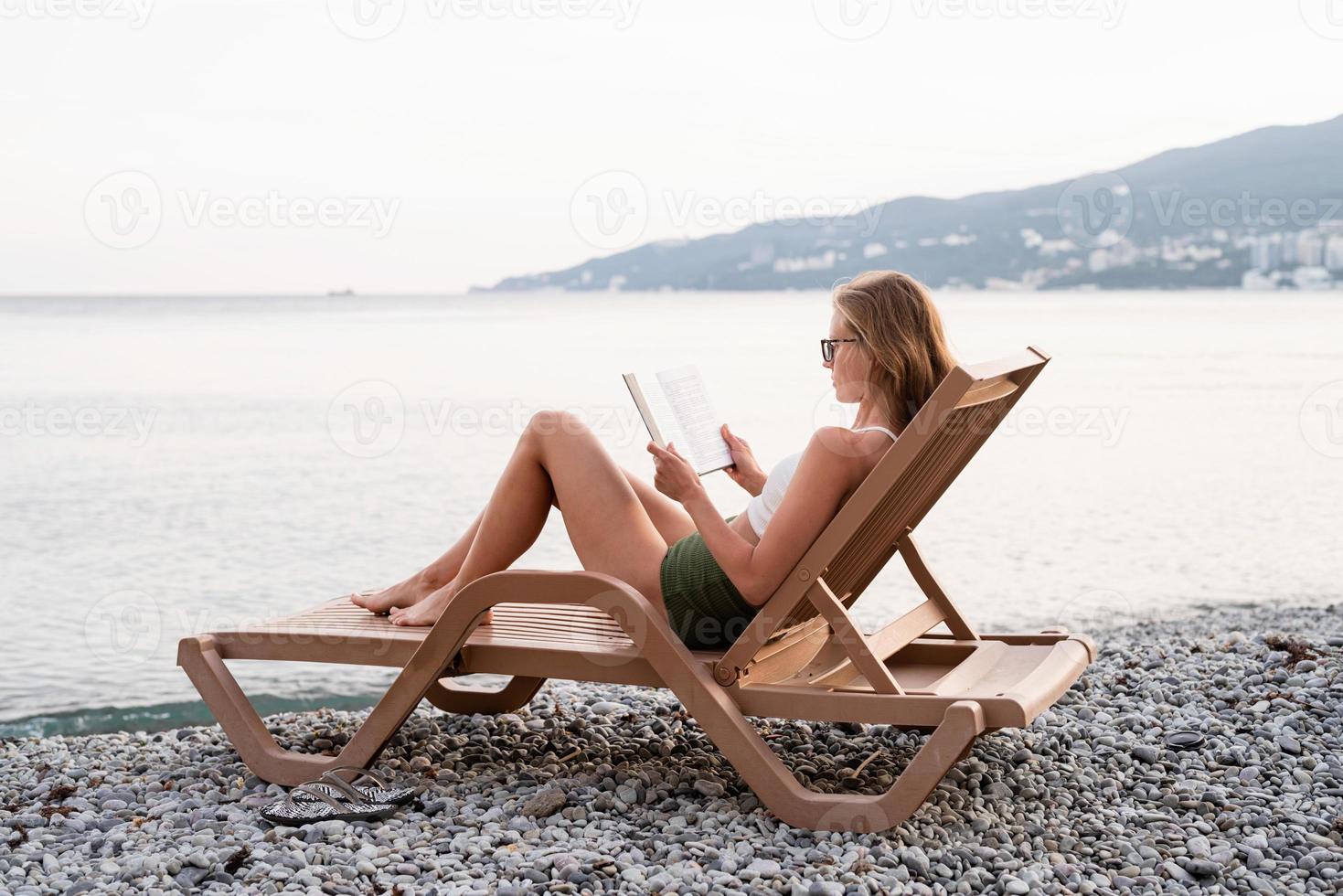 den vackra unga kvinnan som sitter på solstolen och läser en bok foto