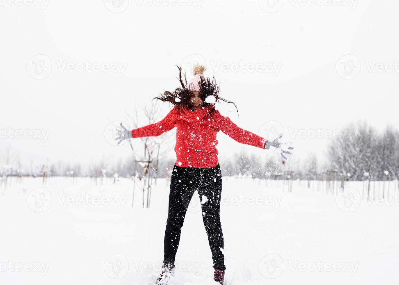 ung brunett sorglös kvinna i röd tröja som hoppar i snö utomhus foto