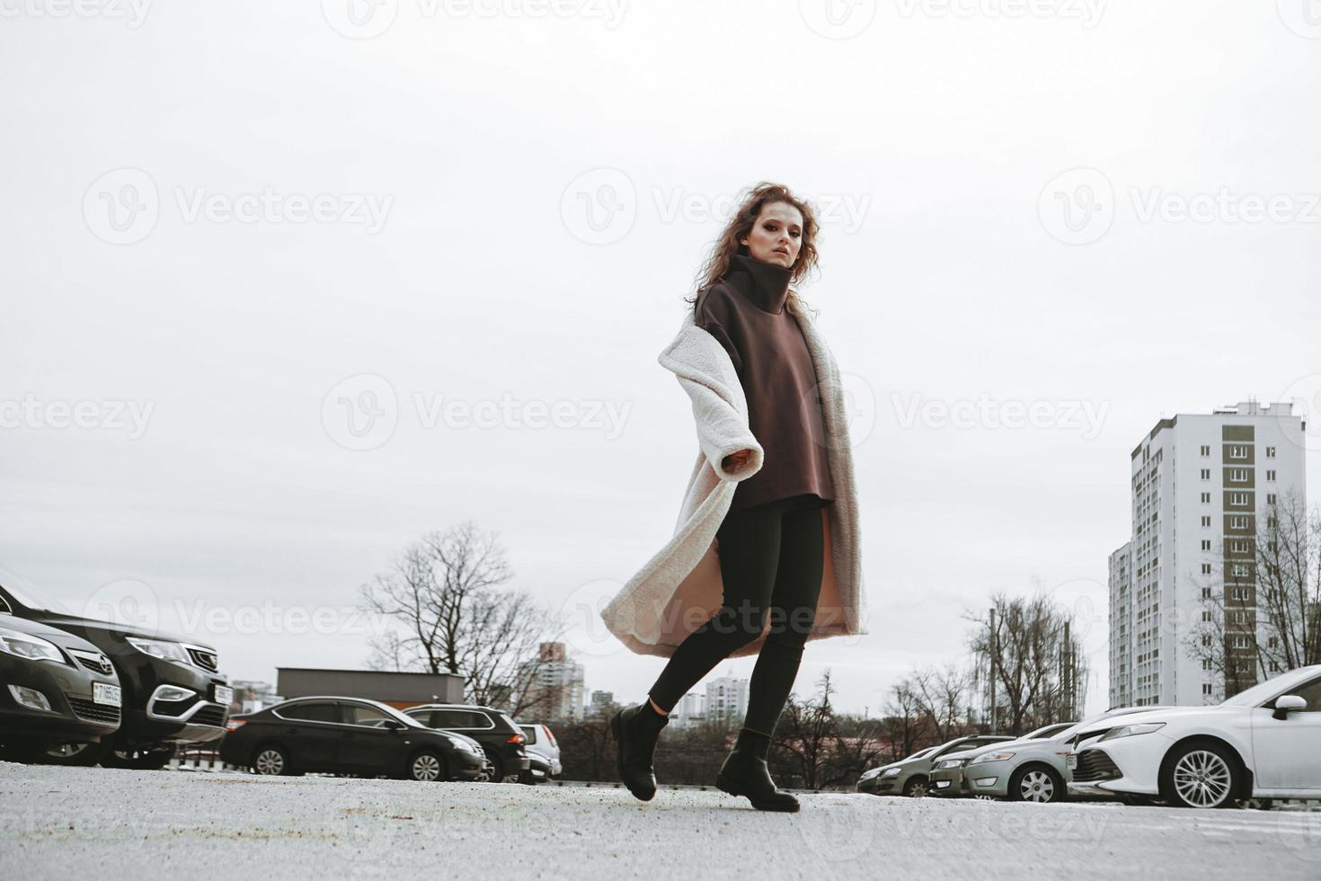 en flicka med rött lockigt hår i vit päls poserar på utomhusparkering foto