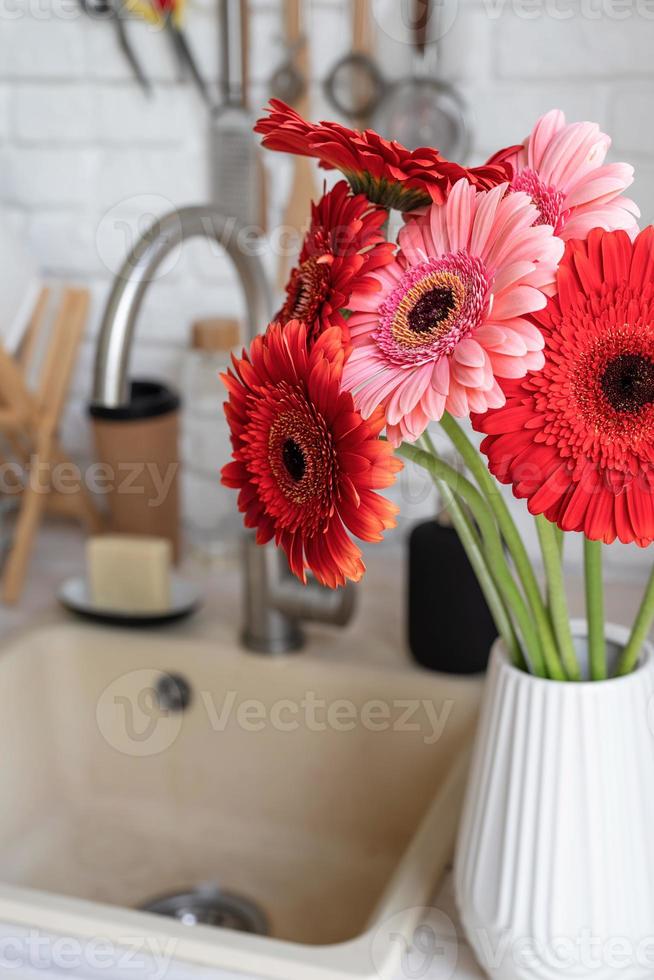 röda och rosa gerbera prästkragar i en vit vas på ett träkök foto