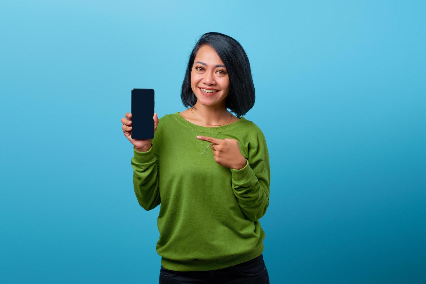 leende asiatisk kvinna som visar tom smartphone -skärm på blå bakgrund foto
