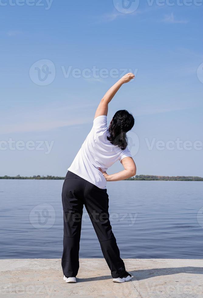 aktiv och glad senior kvinna gör stretching nära floden foto