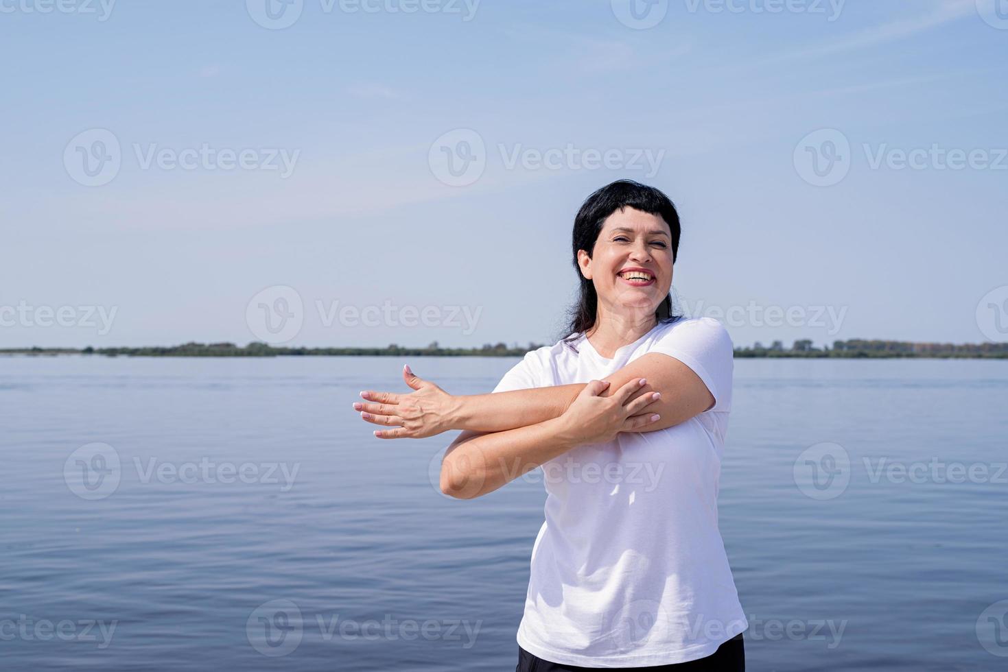 aktiv och glad senior kvinna gör stretching nära floden foto