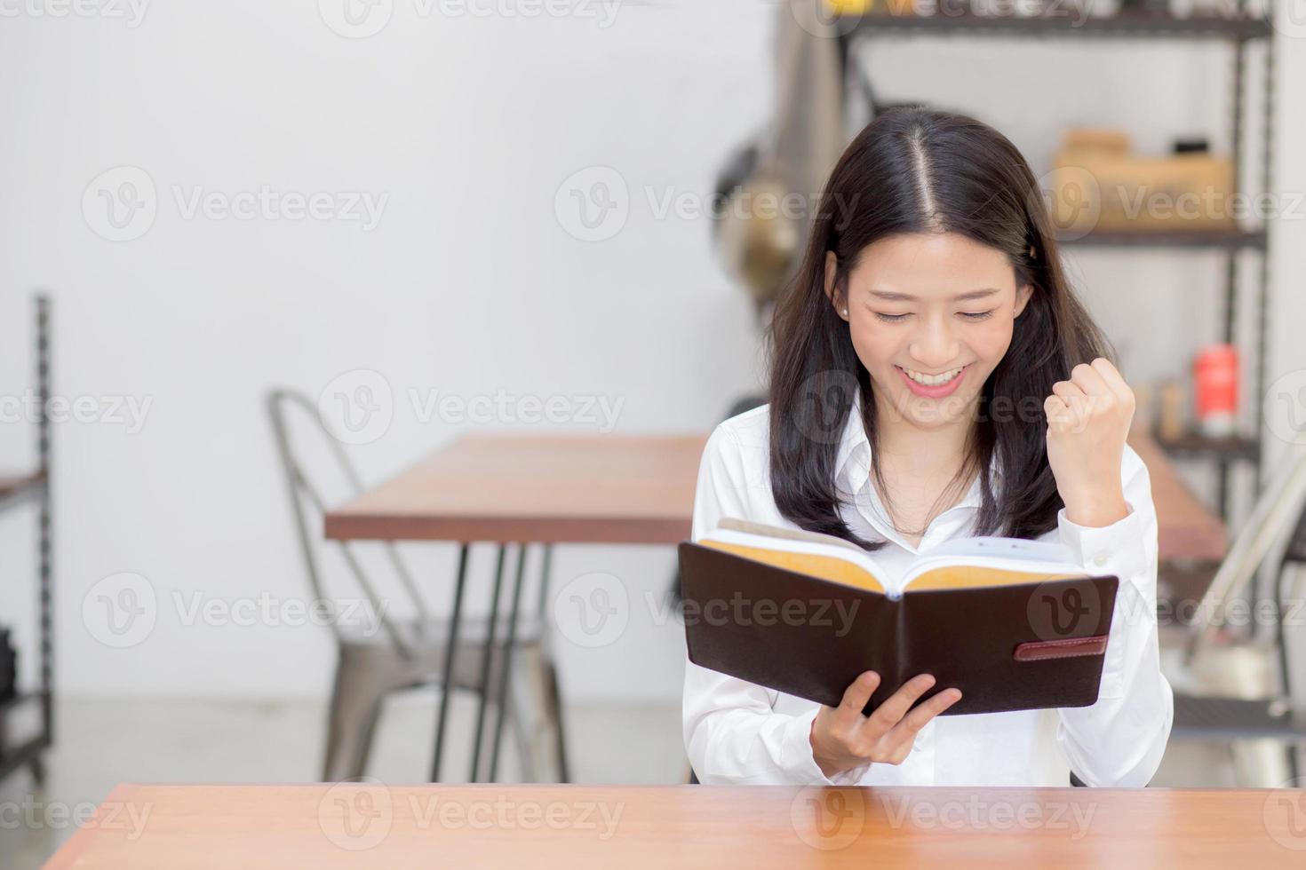vacker av porträtt affärer asiatisk ung kvinna som läser på anteckningsbok med framgång på bordet, flicka tittar anteckning upphetsad på kafé, frilans och utbildning koncept. foto