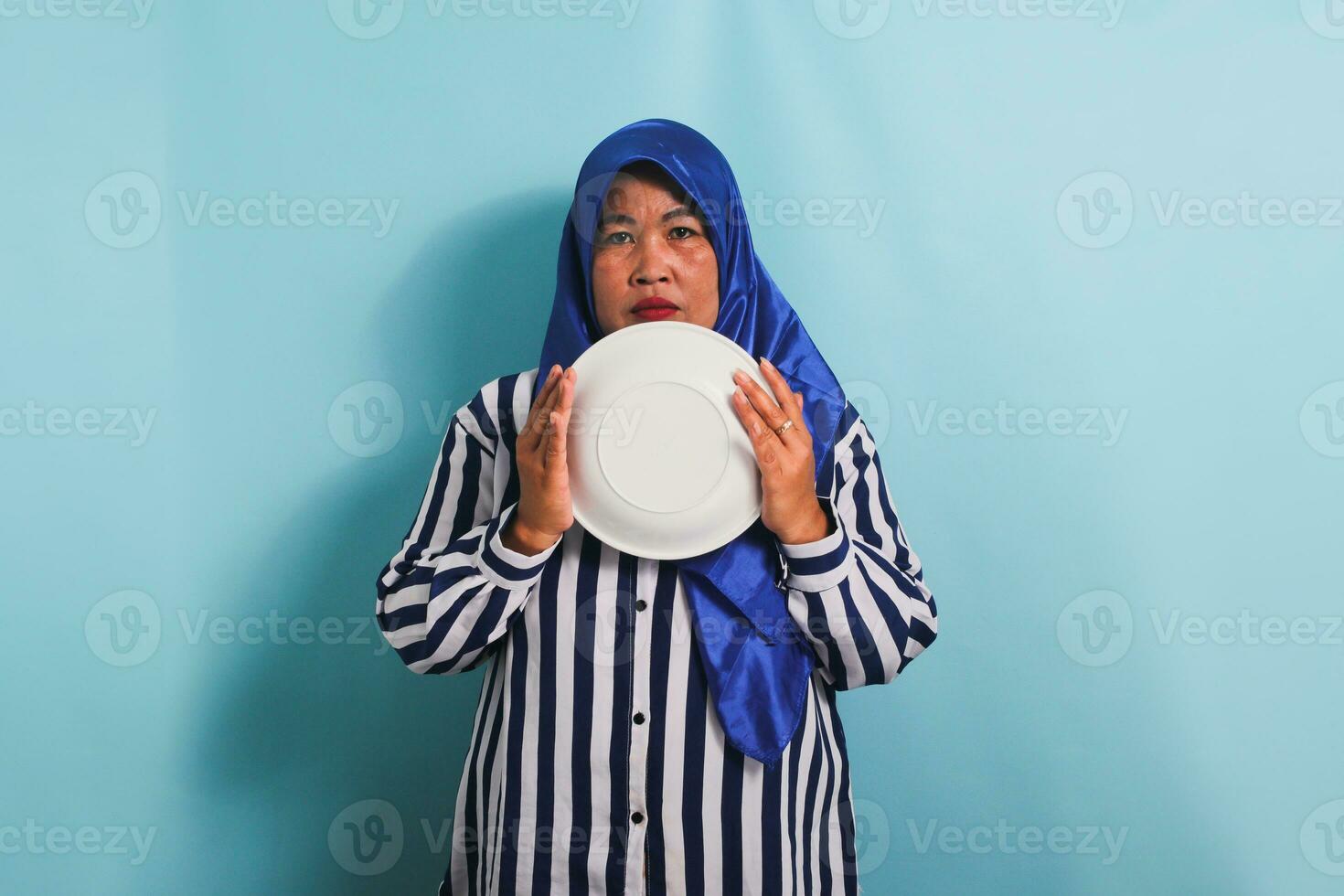 ett olycklig medelålders asiatisk kvinna i en blå hijab och en randig skjorta är innehav ett tömma vit tallrik, känsla hungrig under en diet eller fasta. hon är isolerat på en blå bakgrund foto