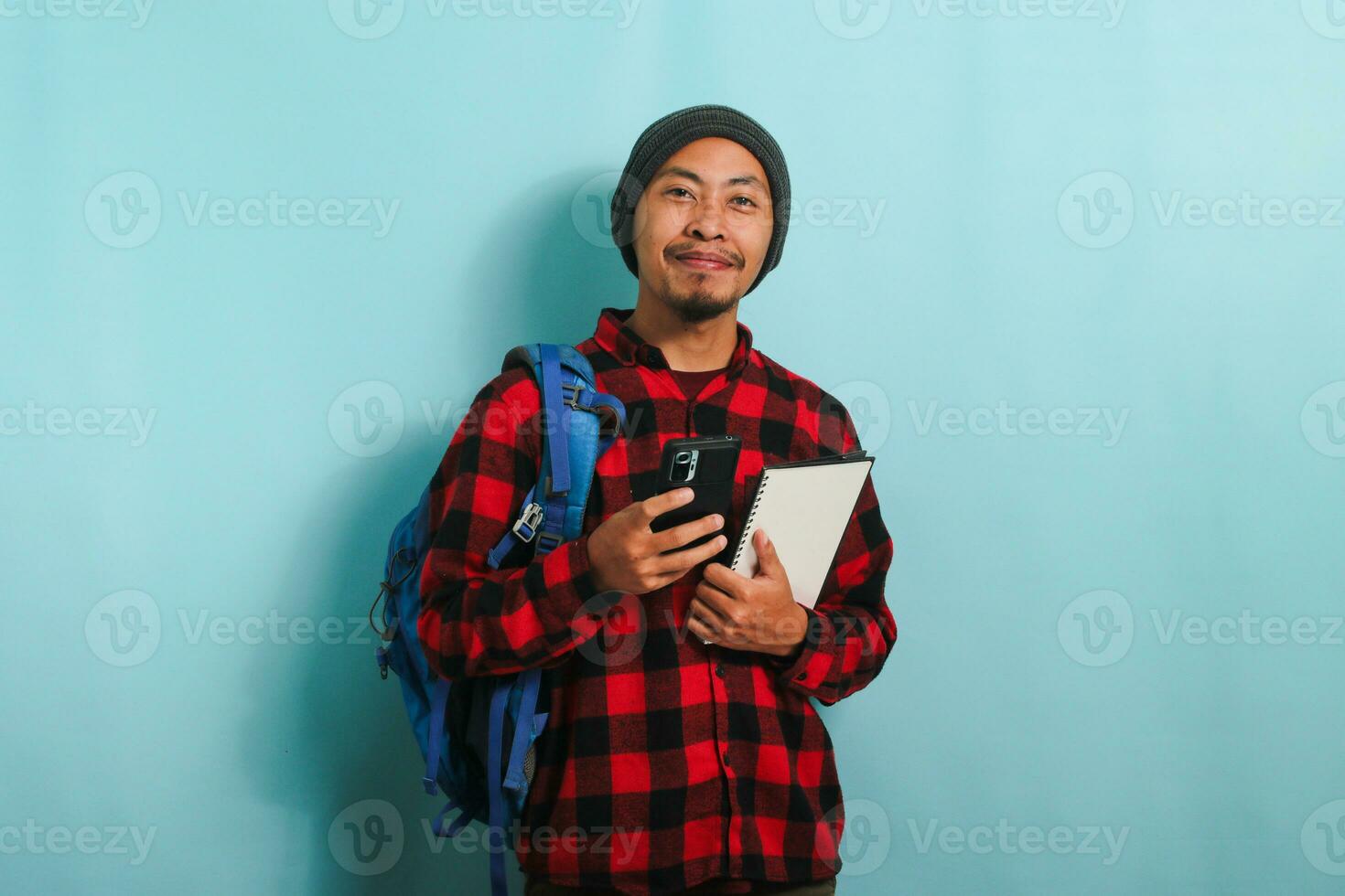 upphetsad ung asiatisk manlig studerande, bär ryggsäck, mössa hatt, och röd pläd flanell skjorta, leende och utseende på kamera, innehar en bok och användningar hans telefon, medan stående mot en blå bakgrund foto