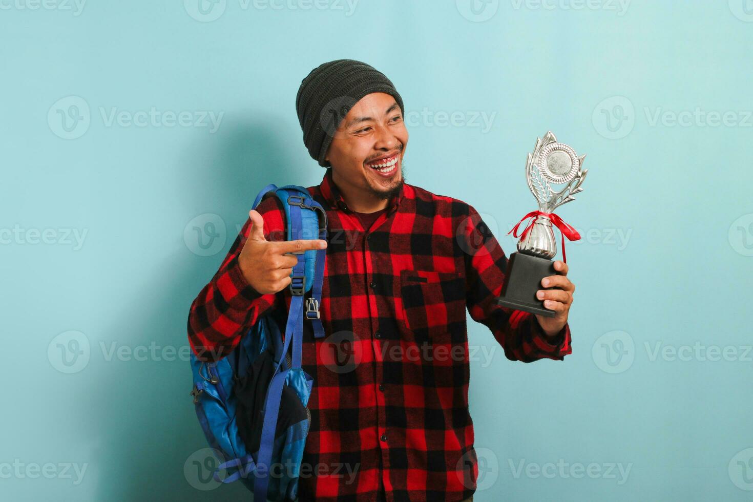 upphetsad ung asiatisk man studerande stolt pekande en trofén i hans hand, isolerat på blå bakgrund foto