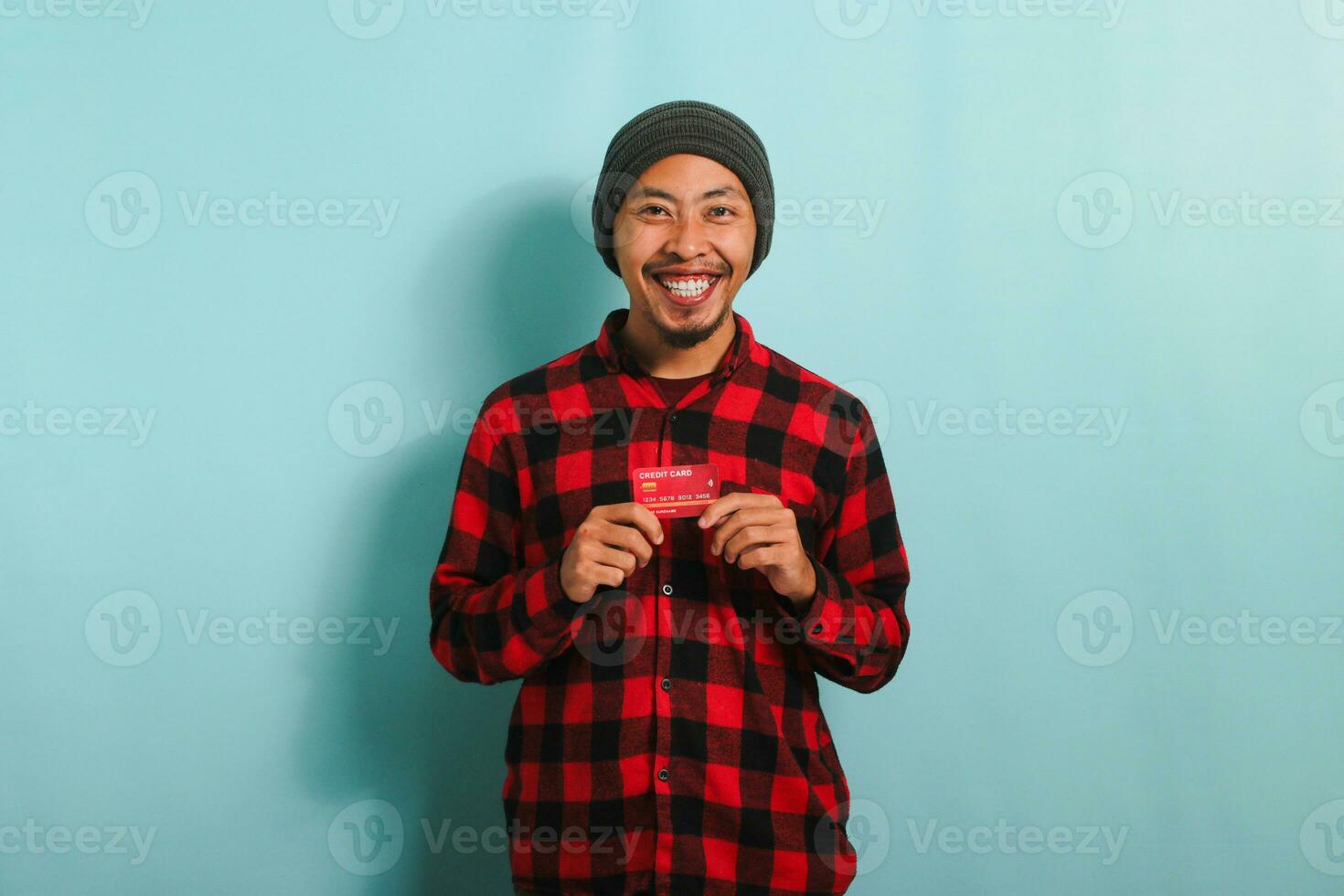 upphetsad ung asiatisk man med en mössa hatt och röd pläd flanell skjorta är innehav Bank kreditera kort i hans händer medan stående mot en blå bakgrund. finansiera, lån, besparingar begrepp foto