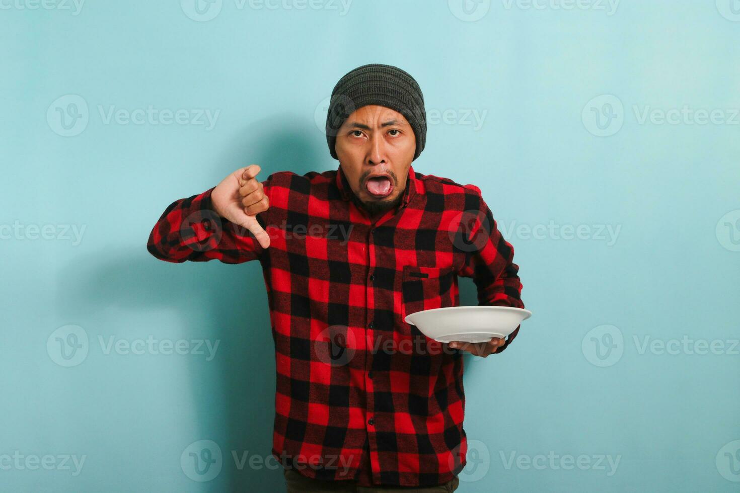 missnöjd ung asiatisk man med en mössa hatt och en röd pläd flanell skjorta är ger en tummen ner, ger de mat en dålig recension, medan innehav ett tömma vit tallrik, isolerat på en blå bakgrund foto