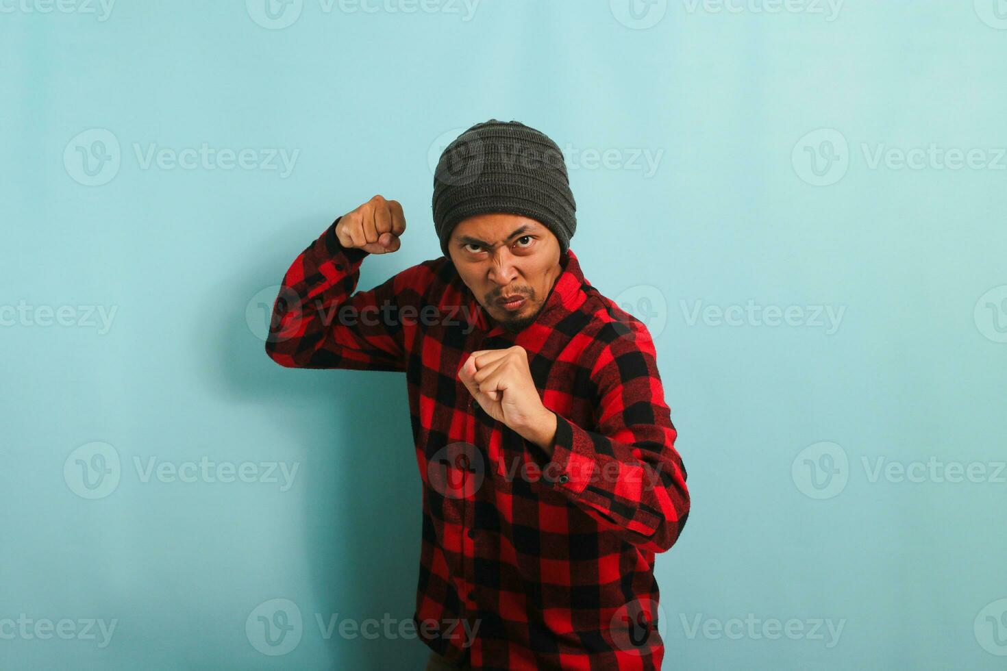ett arg, rasande ung asiatisk man med en mössa hatt och en röd pläd flanell skjorta är redo till bekämpa, med en defensiv näve gest, känsla galen och upprörd. han är isolerat på en blå bakgrund foto