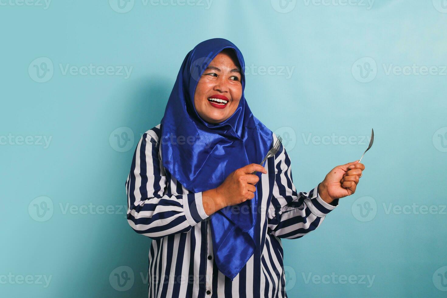 ett upphetsad medelålders asiatisk kvinna i en blå hijab och en randig skjorta är pekande till de rätt copy med en sked och gaffel. hon är isolerat på en blå bakgrund foto