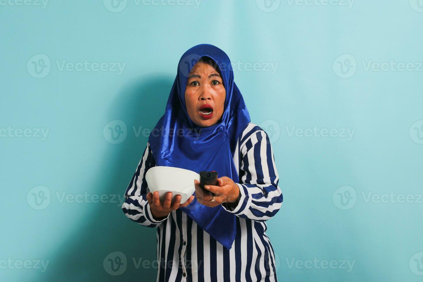 en chockade medelålders asiatisk kvinna i en blå hijab är tittar på tv, pekande de TV avlägsen kontrollant mot de kamera, och växlande kanaler med ett öppen mun, isolerat på en blå bakgrund foto