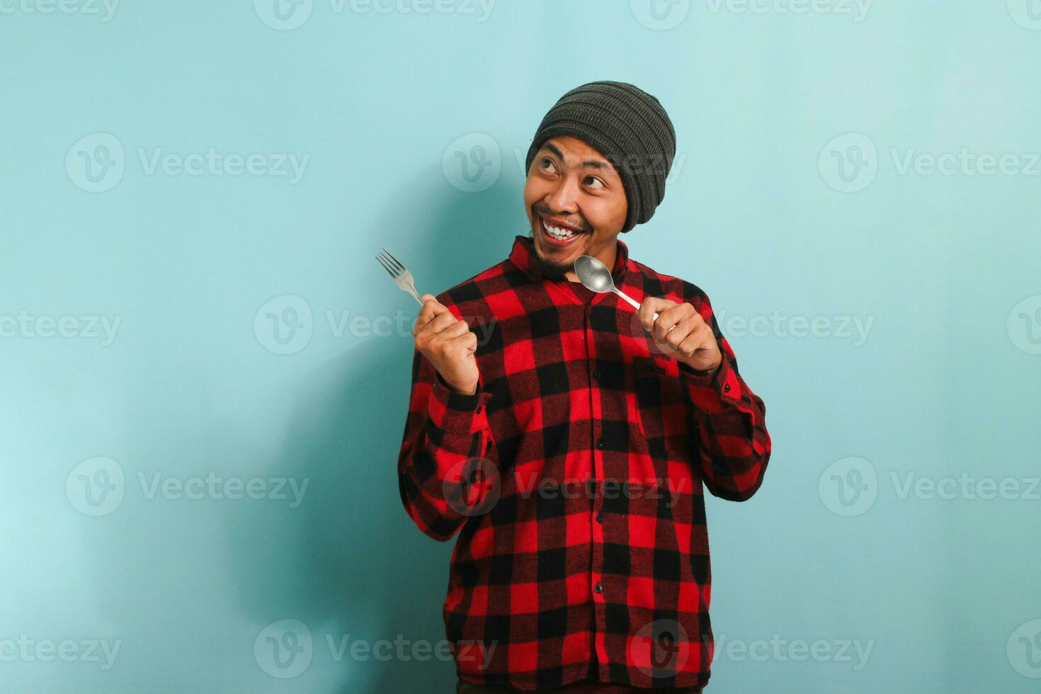 upphetsad ung asiatisk man med mössa hatt och röd pläd flanell skjorta pekande åt sidan på kopia Plats med en sked och gaffel, som om indikerar en frestande måltid eller mat alternativ, isolerat på en blå bakgrund foto