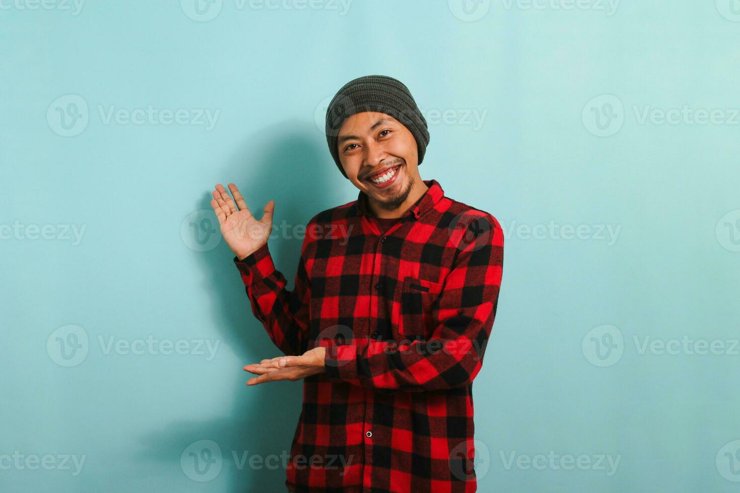 ett upphetsad ung asiatisk man med en mössa hatt och en röd pläd flanell skjorta är som visar med hans händer till de sida, indikerar de tömma kopia Plats, isolerat på en blå bakgrund foto