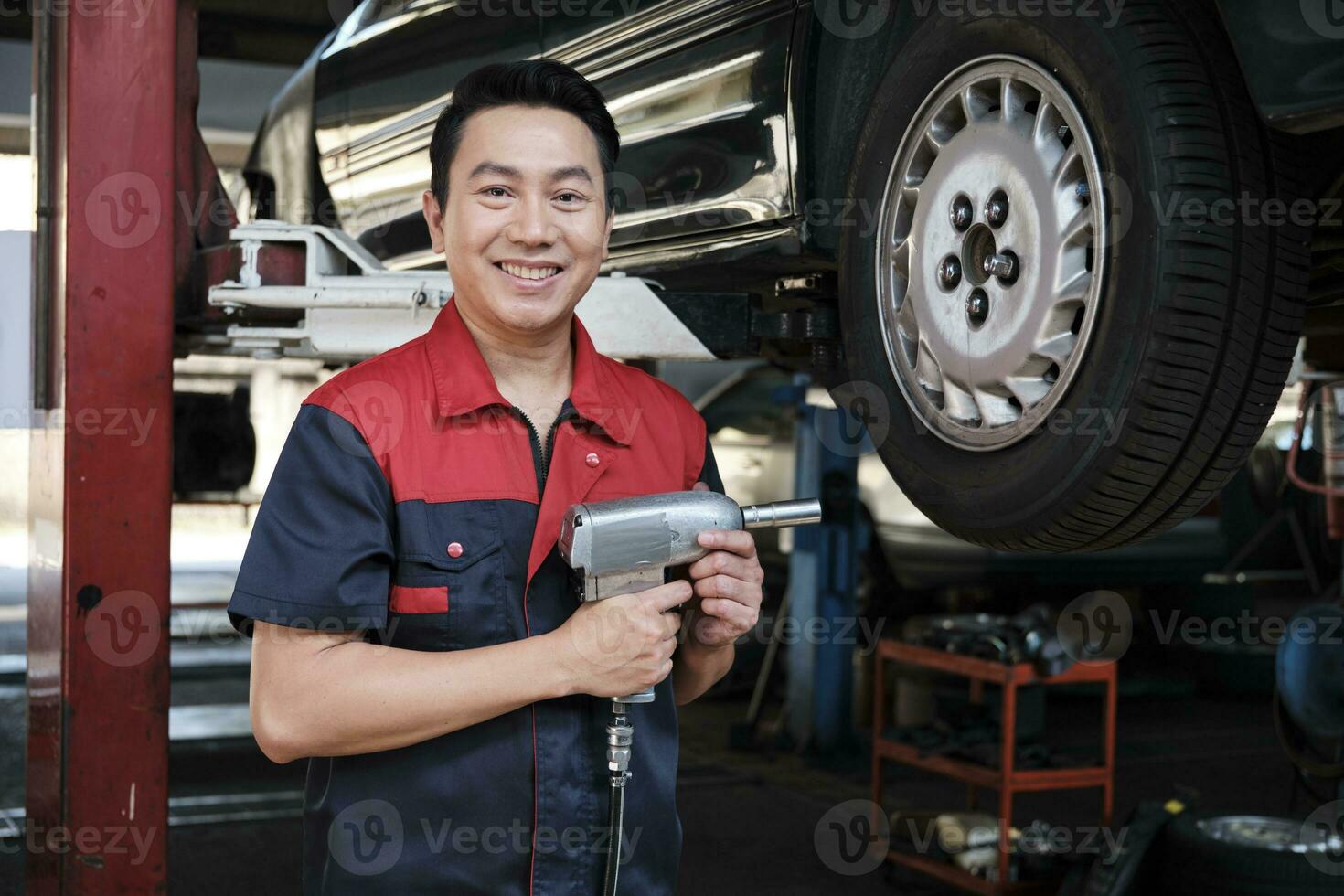 ett expert- asiatisk manlig bil- mekaniker tekniker är skruvning bil hjul nötter på lyft med borra rycka för reparera på garage. fordon underhåll service Arbetar industri ockupation företag jobb. foto