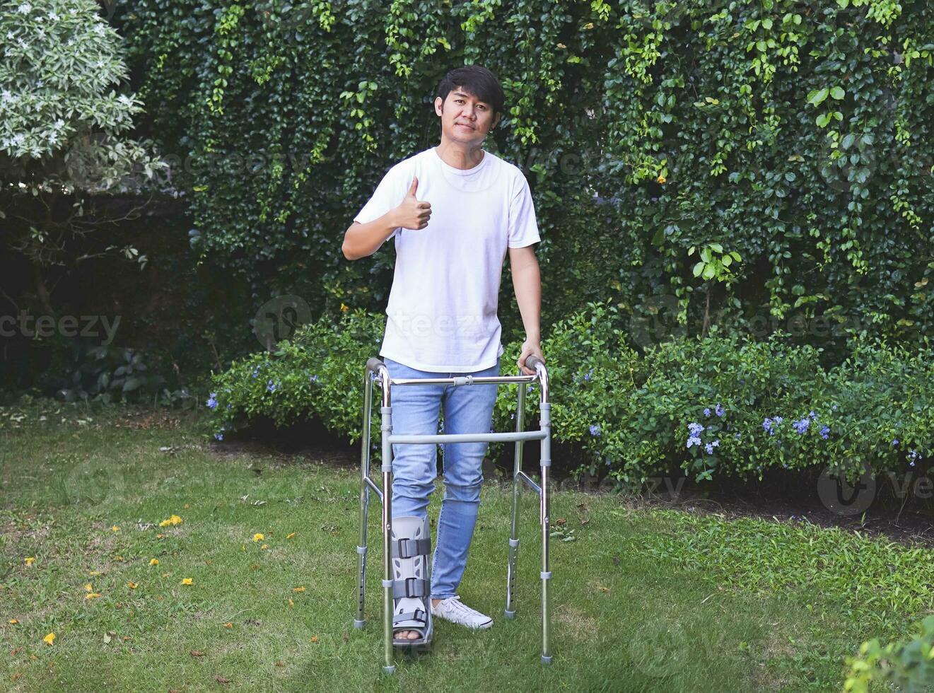 asiatisk ung man bär luftsändning känga gående i de trädgård med ortopedisk rollator. som visar tumme upp och ser på kamera. foto