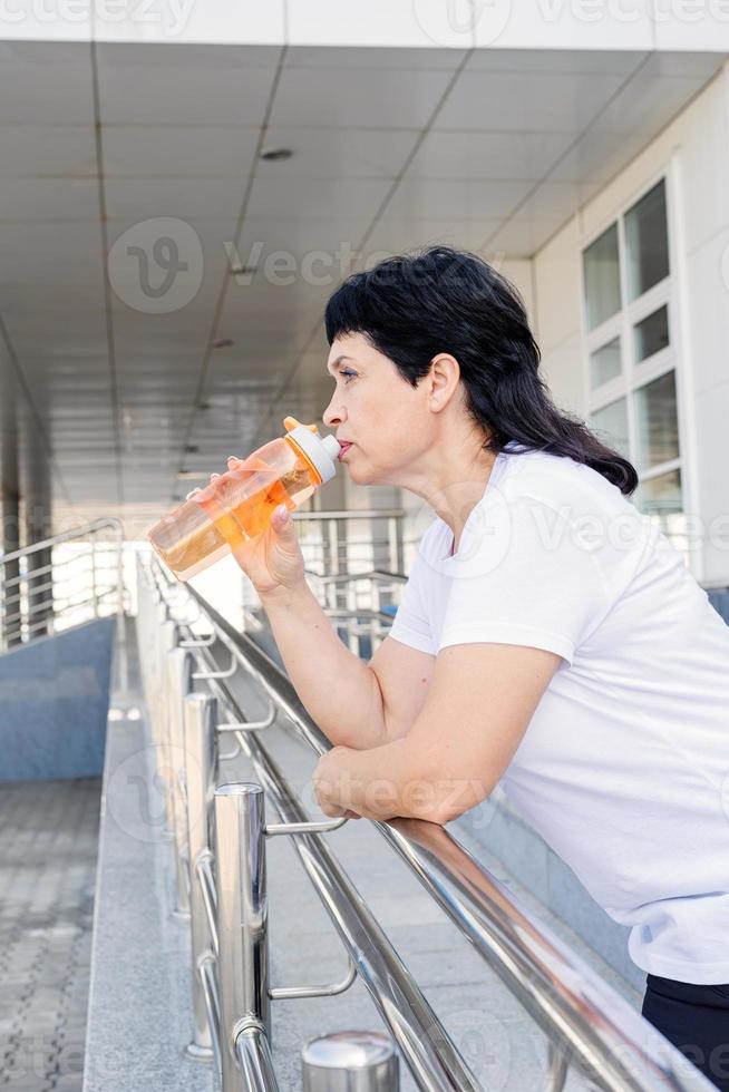 senior kvinna som dricker vatten efter träning utomhus på urban bakgrund foto
