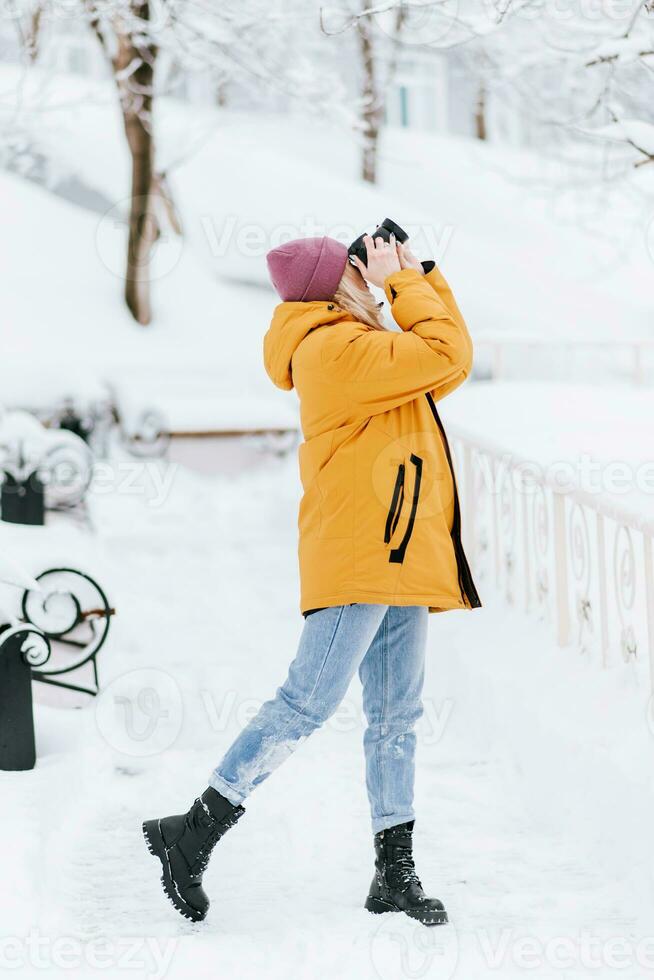 skön flicka i en gul jacka fotograf tar bilder av snö i en vinter- parkera foto