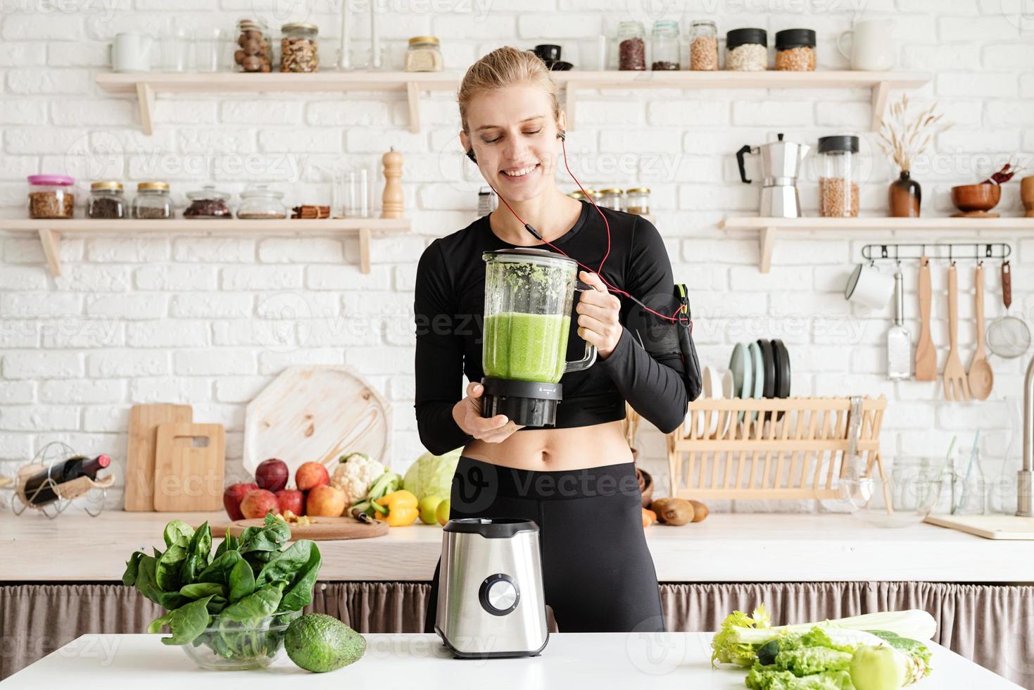 ung blond leende kvinna som gör grön smoothie hemma i köket foto