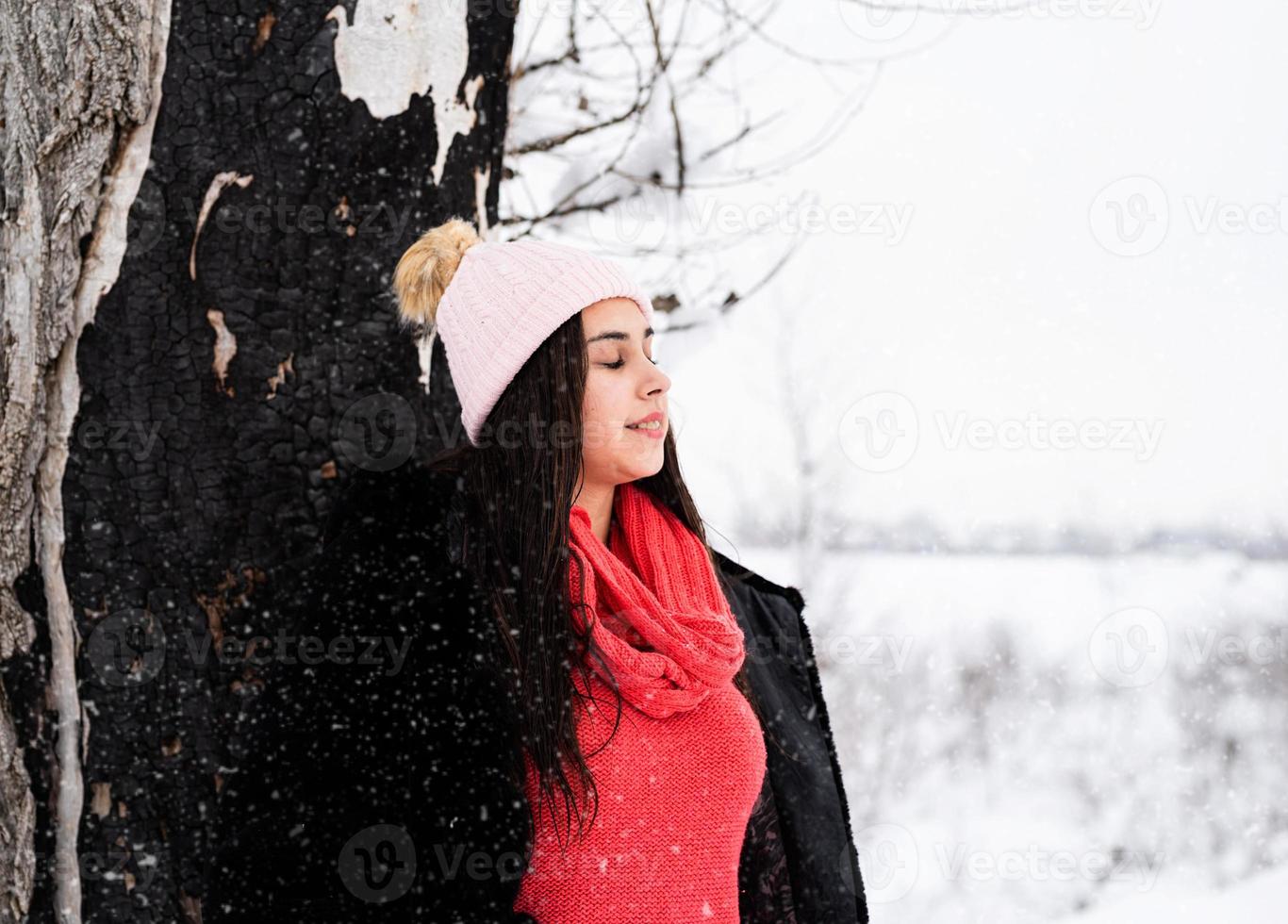 ung kvinna som står vid trädet med ögonen stängda i snöfall foto