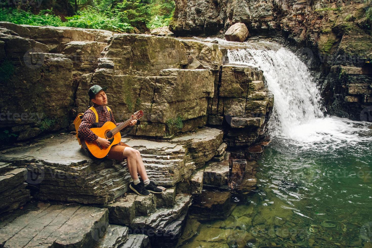 man spelar gitarr som sitter vid ett vattenfall foto