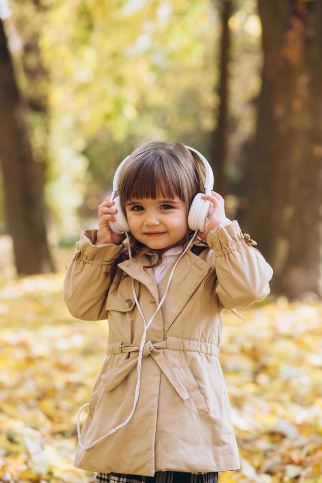 glad liten flicka som lyssnar på musik på hörlurar i höstparken. foto