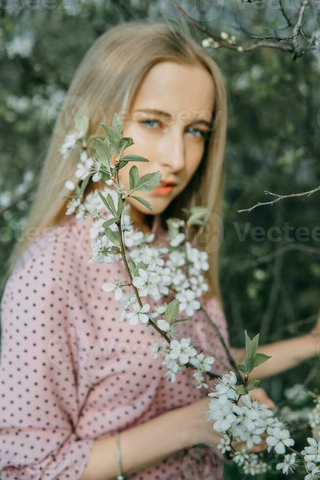 blond flicka på en vår promenad i de trädgård med körsbär blommar. kvinna porträtt, närbild. en flicka i en rosa polka punkt klänning. foto