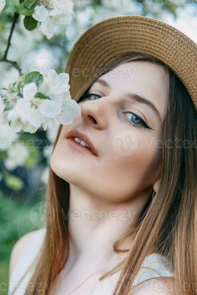 skön ung flicka i vit klänning och hatt i blomning äpple fruktträdgård. blomning äpple träd med vit blommor. foto