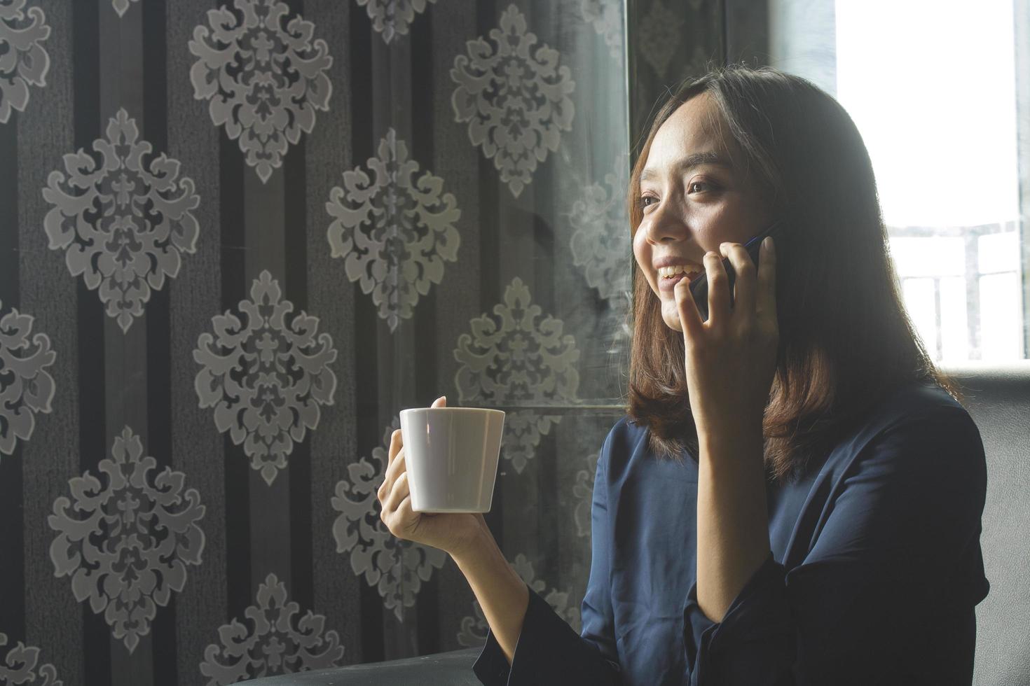 avslappnad leende asiatisk kvinna som dricker kaffe medan hon ringde om jobbet foto