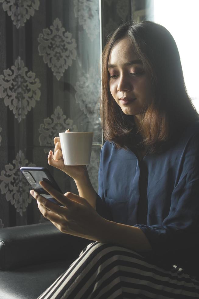 avslappnad asiatisk kvinna som dricker kaffe medan hon använder en mobiltelefon för arbete foto