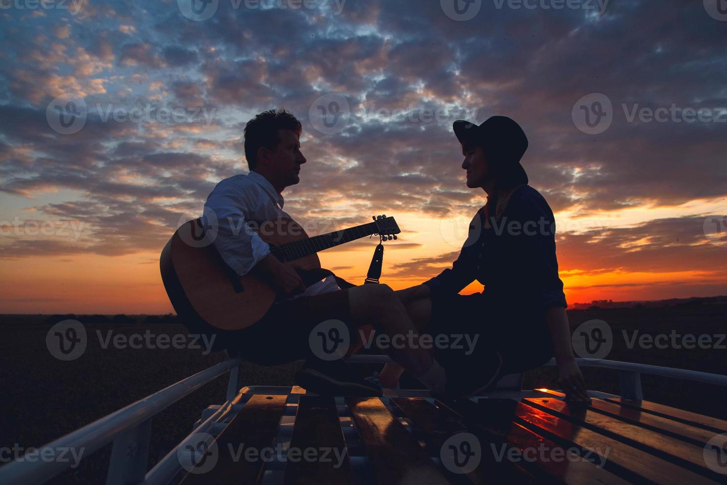 silhuett av man med gitarr och kvinna på taket av en bil vid solnedgången foto