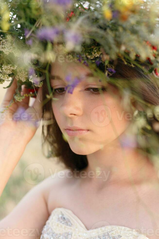 skön kvinna med en krans på henne huvud Sammanträde i en fält i blommor foto