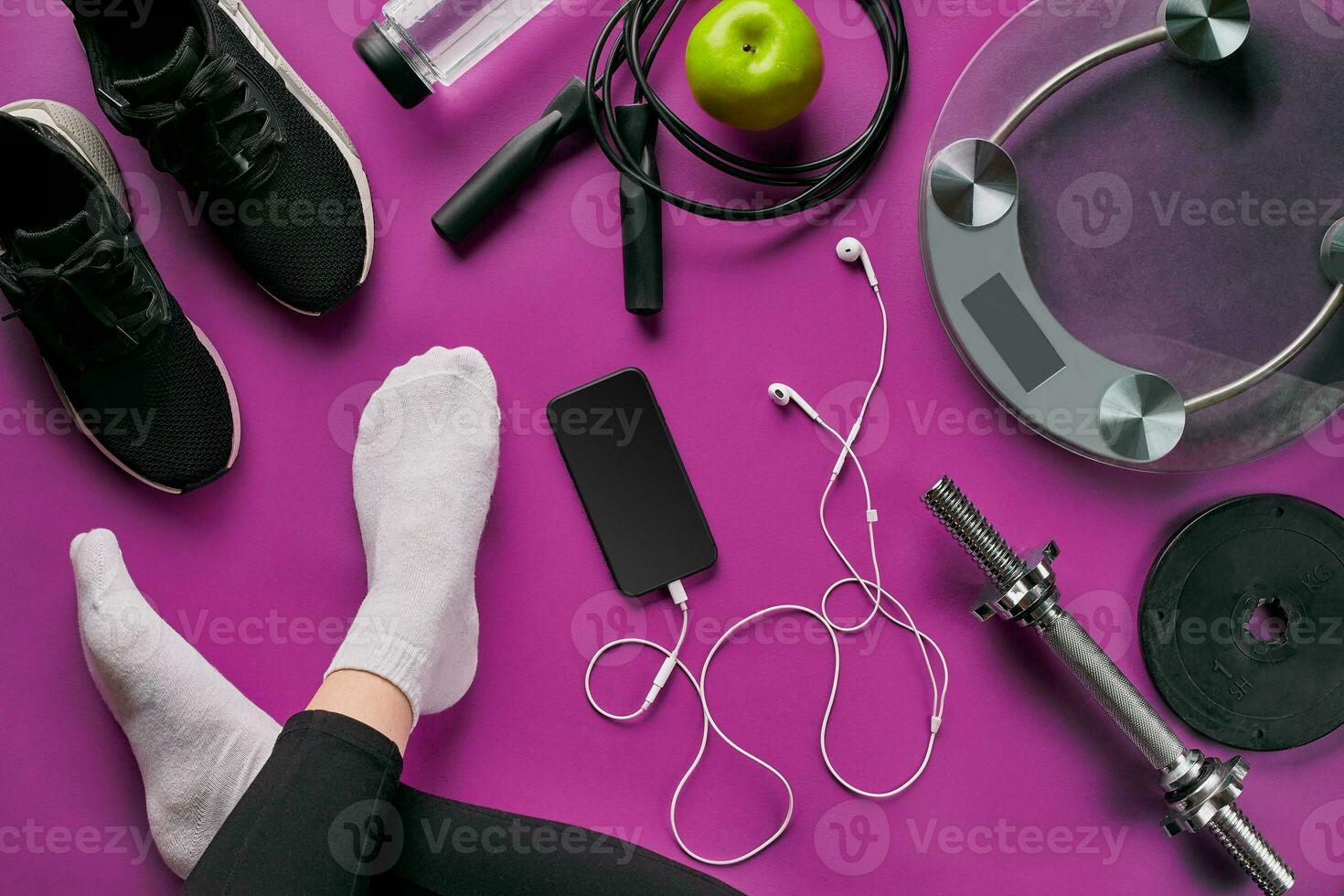 platt lägga Gym Utrustning sådan som hoppa rep, flaska av vattnen, smartphone med hörlurar, äpple och mänsklig ben i strumpor på Färg bakgrund. foto