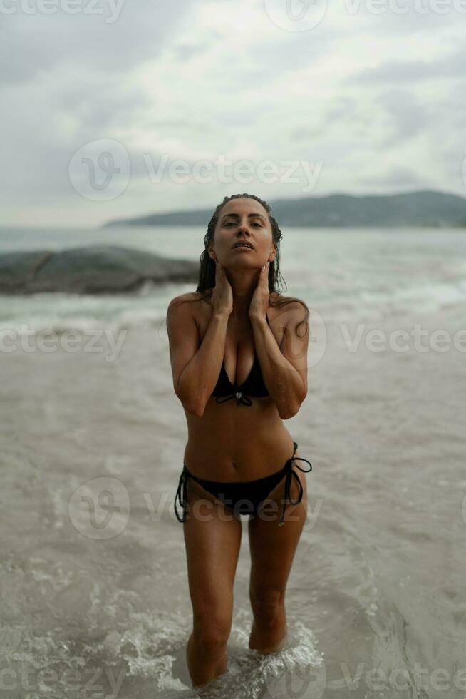 underbar brunett kvinna med perfekt figur Framställ på tropisk strand. bär eleganta svart badkläder. foto