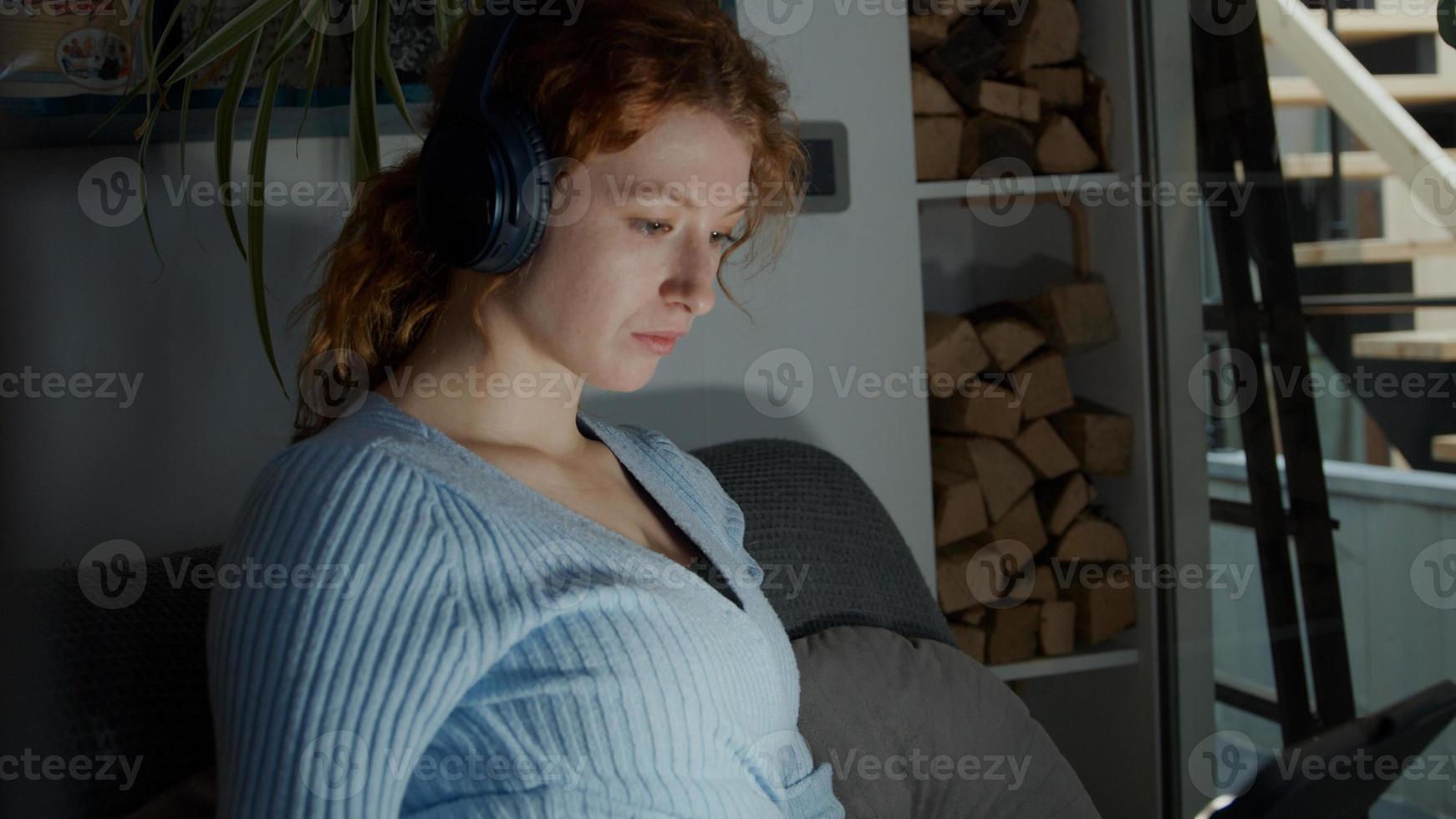 ung vit kvinna i vardagsrummet, hörlurar på öronen som tittar på surfplattan foto
