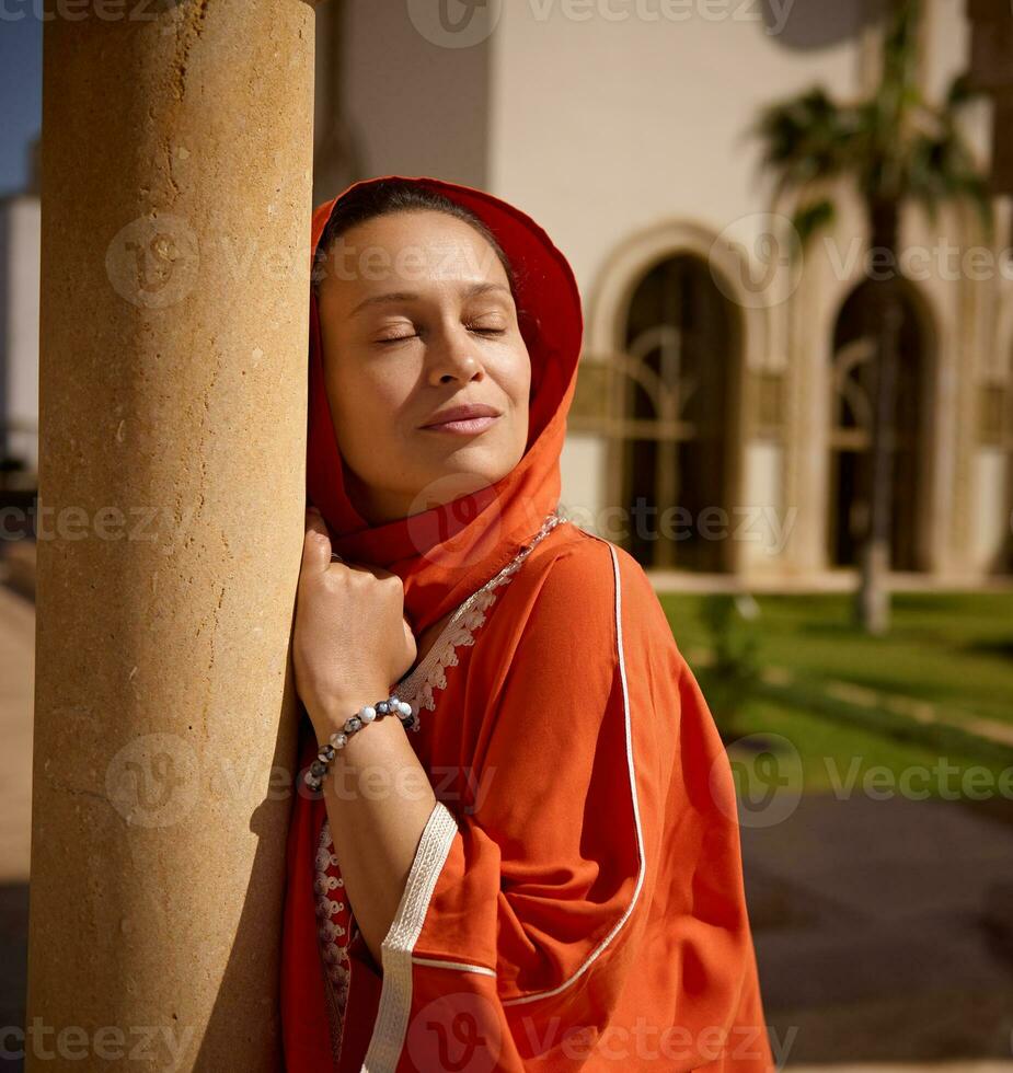 underbar muslim kvinna med henne ögon stängd, bär ljus orange slöja och traditionell kläder på hassan ii moské foto