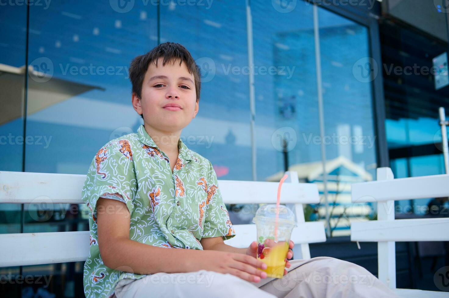 stilig pre tonåring pojke i sommar skjorta, innehav en glas av fräsch pressas orange juice, självsäkert ser på kamera foto