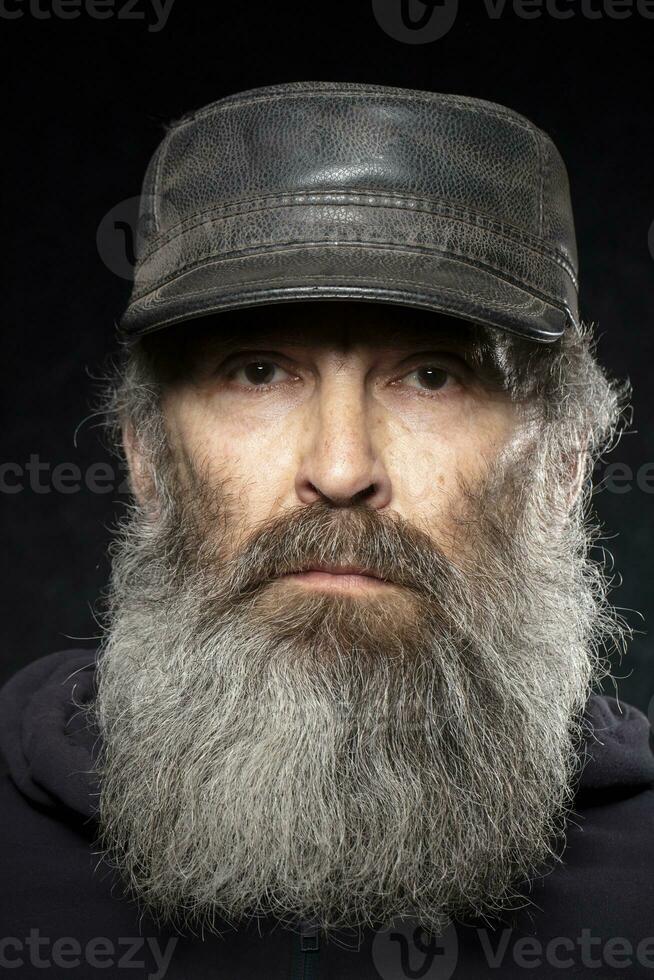 de ansikte av ett äldre man med en grå skägg i en läder hatt på en svart bakgrund. foto