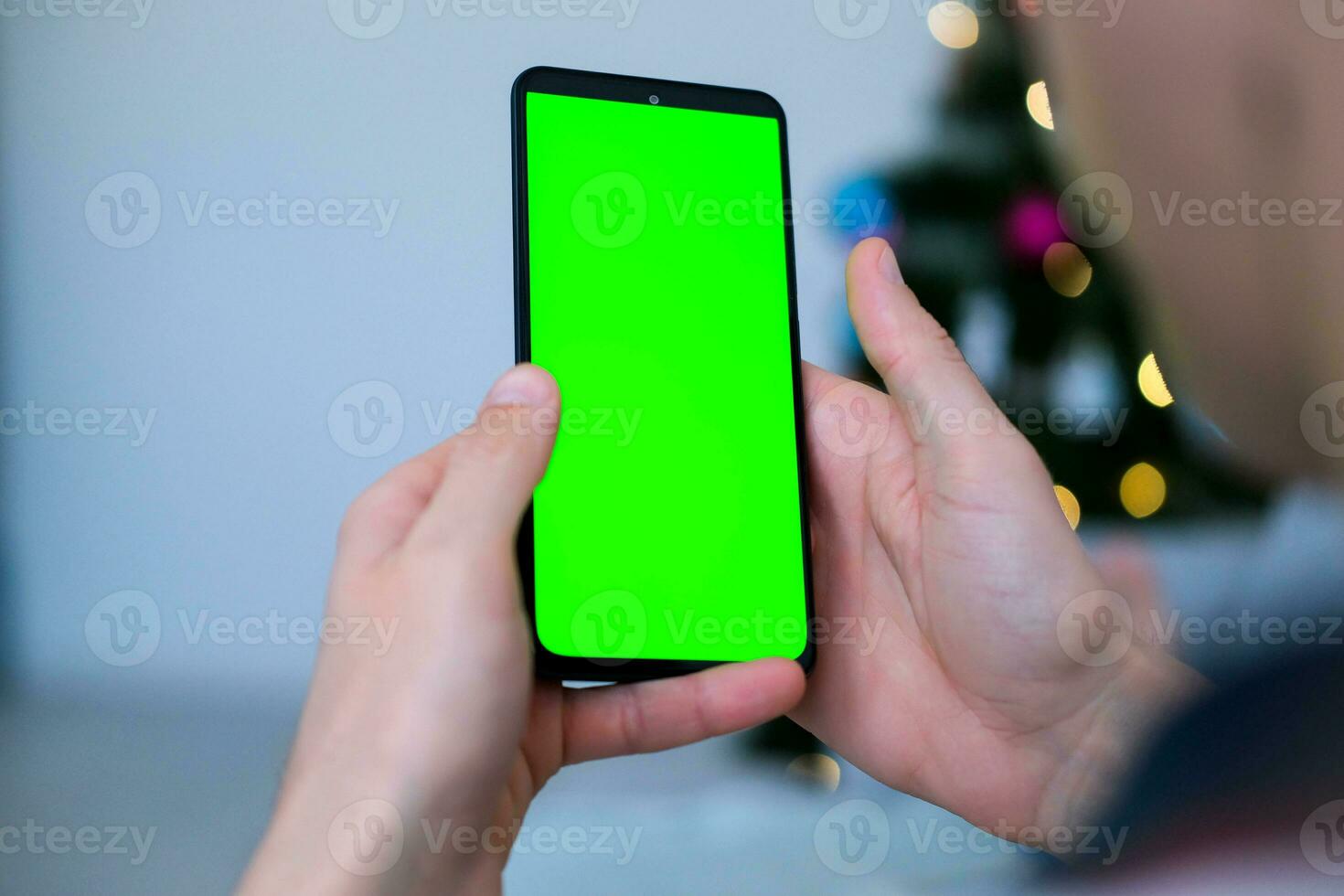 närbild skott av man hand använder sig av mobil telefon med grön skärm mot de bakgrund av en jul träd. finger svep upp och ner i grön skärm av de mobil. grön skärm attrapp mall . foto