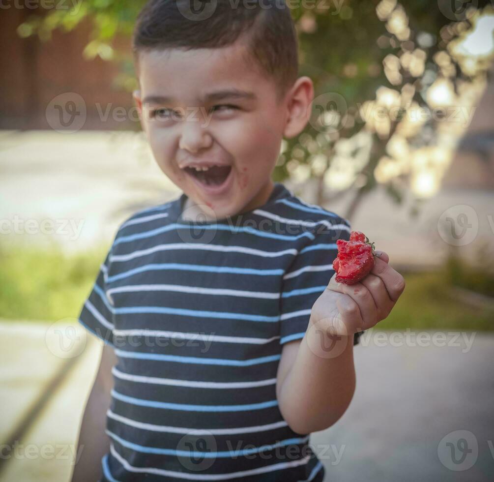 en liten pojke äter jordgubbar. sommar mat. en ung unge äter en smaskigt jordgubb i varm sommar dag foto