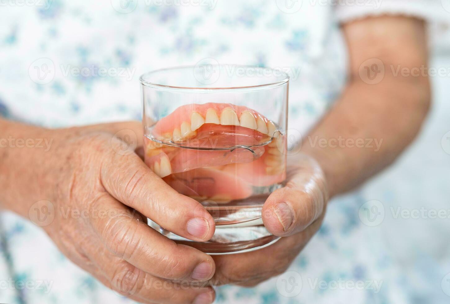 asiatisk senior kvinna patient innehav och tvättning tandprotes i vatten rengöringsmedel glas för Bra tugga. foto