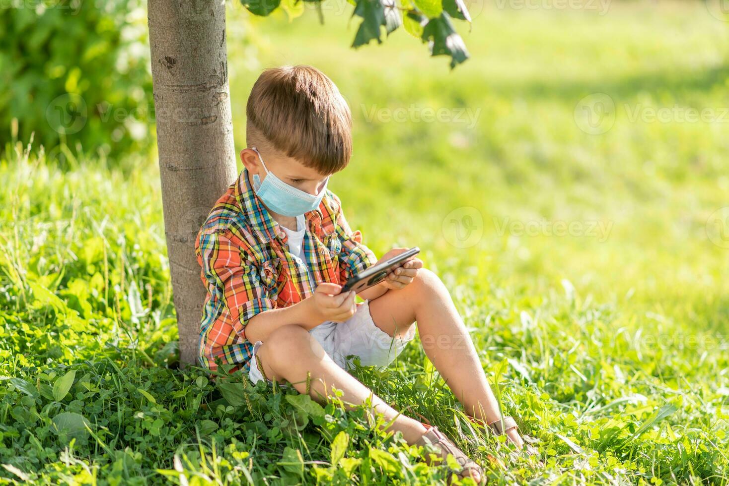 en unge i en medicinsk mask sitter på de gräs och utseende i de telefon tecknade serier i de sommar på solnedgång. barn med en mobil telefon i hans händer. förebyggande mot coronavirus covid-19 under en pandemi foto