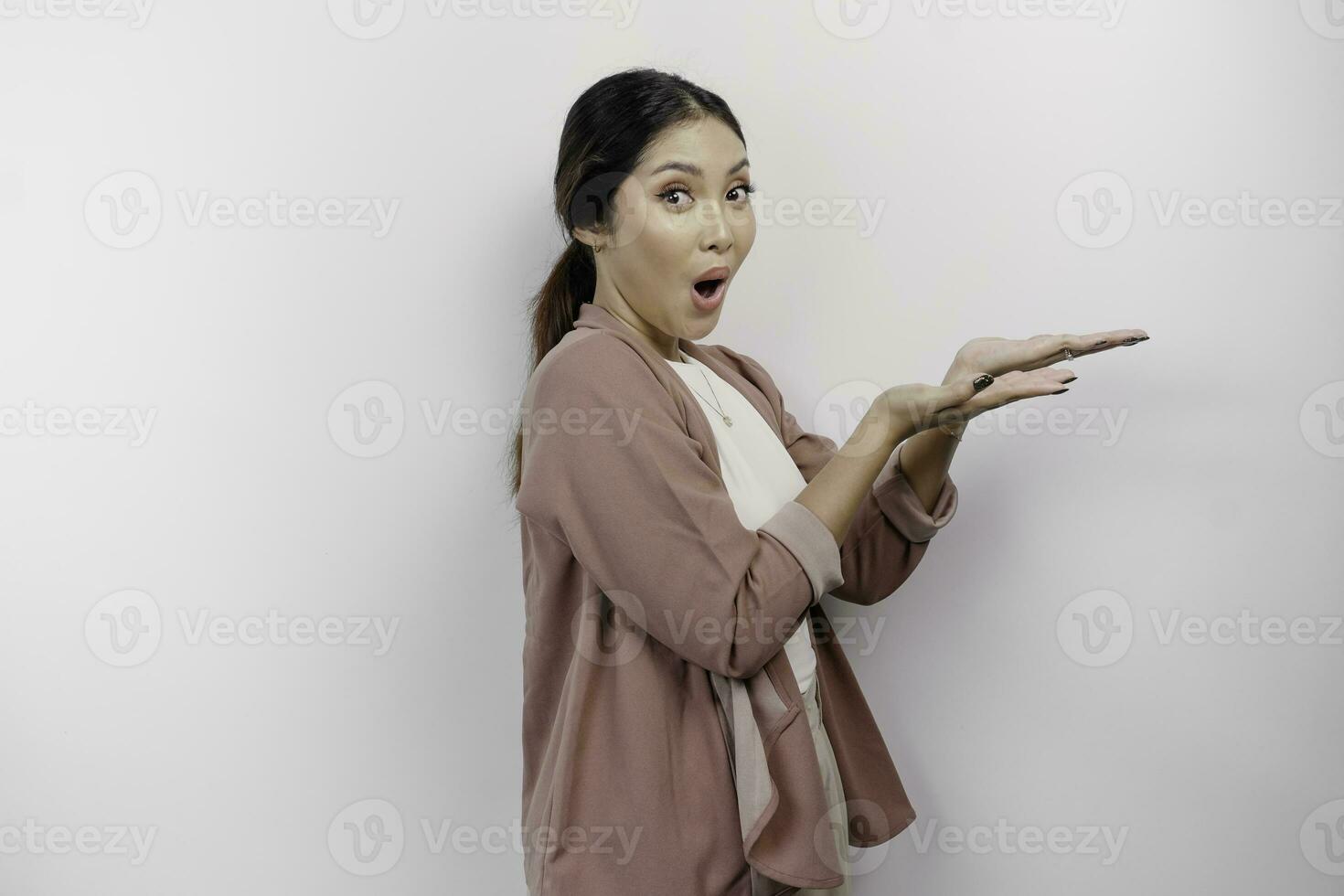 chockade asiatisk kvinna anställd bär en cardigan pekande på de kopia Plats bredvid henne, isolerat förbi en vit bakgrund foto