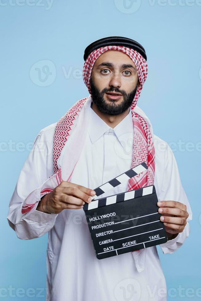 film scen direktör bär traditionell muslim kläder innehav clapperboard och ser på kamera. arab klädd i islamic slöja och thobe använder sig av clapboard studio porträtt foto