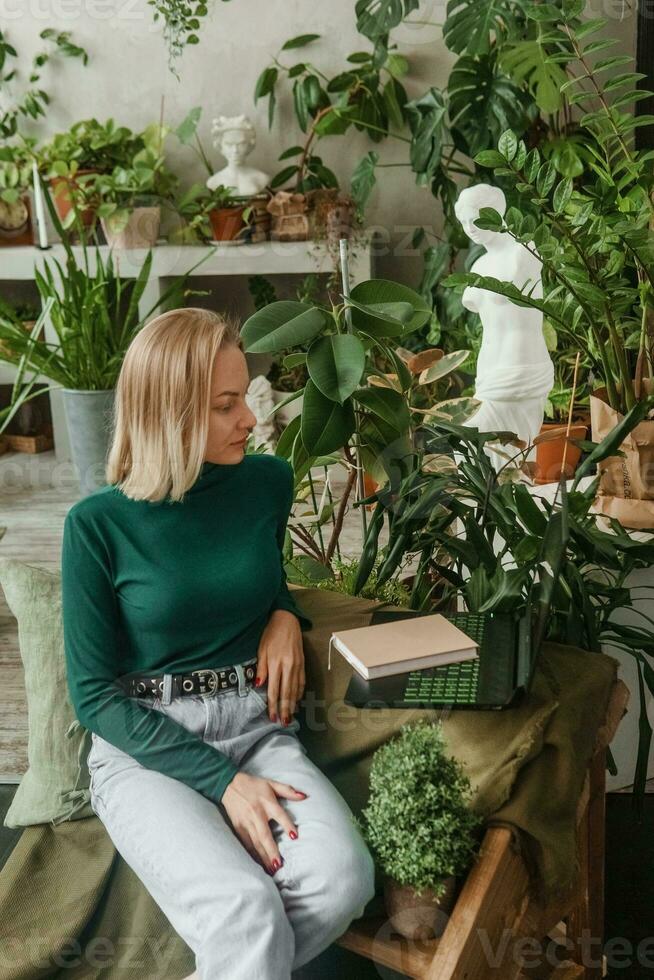 en blond kvinna i en rum med en massa av grön inomhus- växter är arbetssätt på en bärbar dator. de begrepp av biofysiska design i de interiör. arbete från Hem, arbete som en frilansare. foto