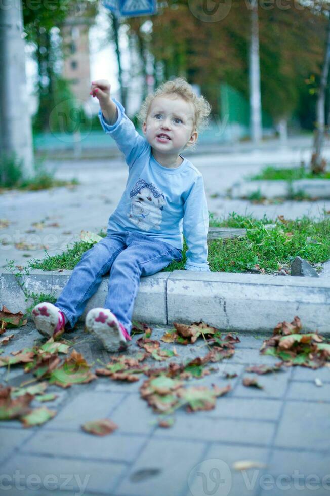 liten lockigt flicka med blond hår Sammanträde på de trottoarkant. en barn poäng till något förbi hand. foto