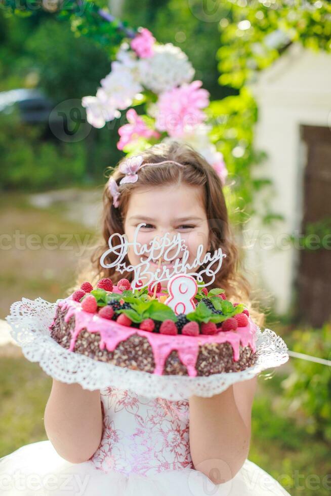 flicka innehav födelsedag kaka med siffra 8 och inskrift Lycklig födelsedag. fira ceremoniell evenemang på gata under karantän. barn i bakgrund dekorerad Foto zon av blommor.