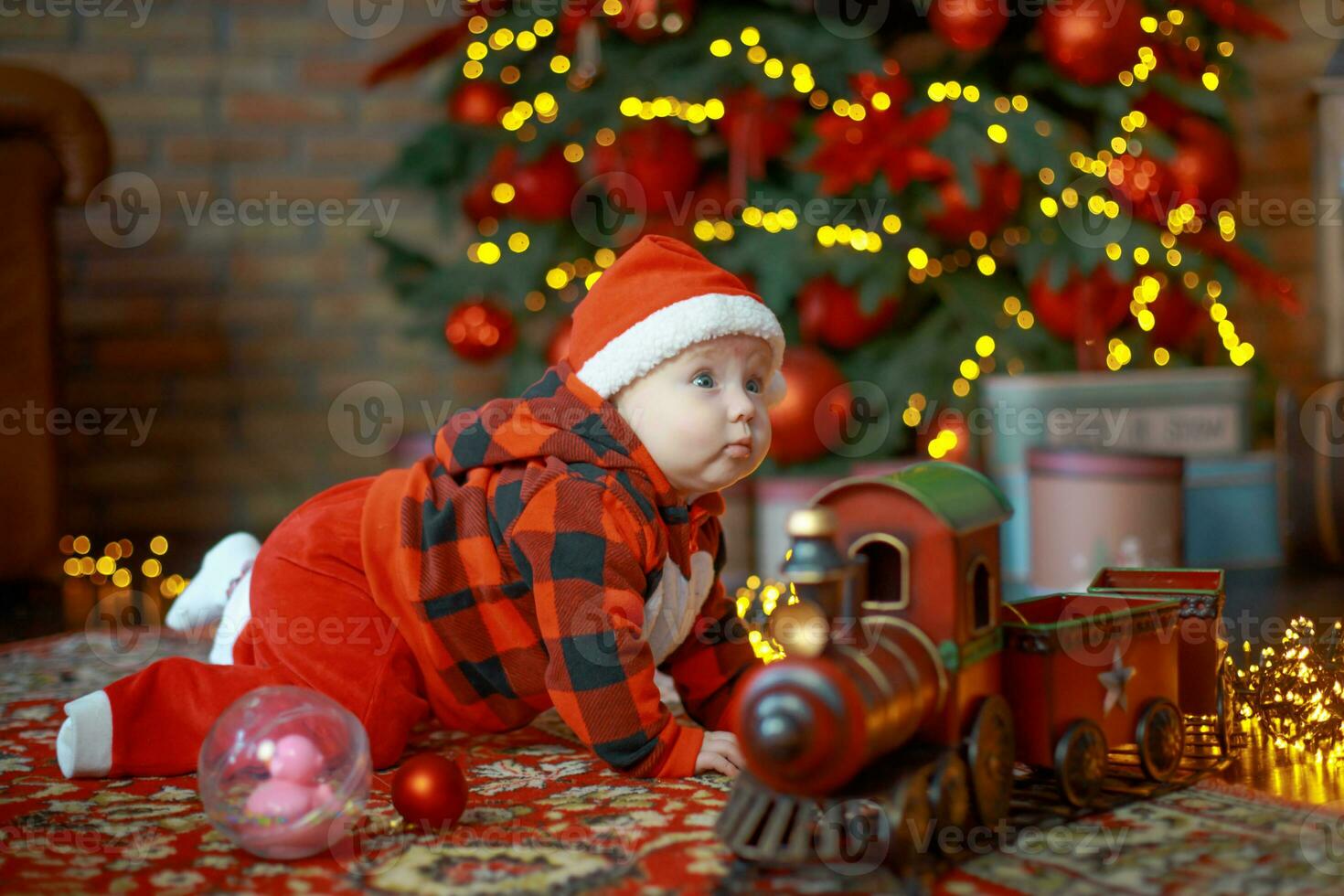 liten flicka Nästa till jul tåg. Lycklig barn i santa hatt nära gåvor på bakgrund av ny år träd i festlig rum. 6 månad gammal bebis är leende. skön liten flicka väntar för jul presenterar. foto