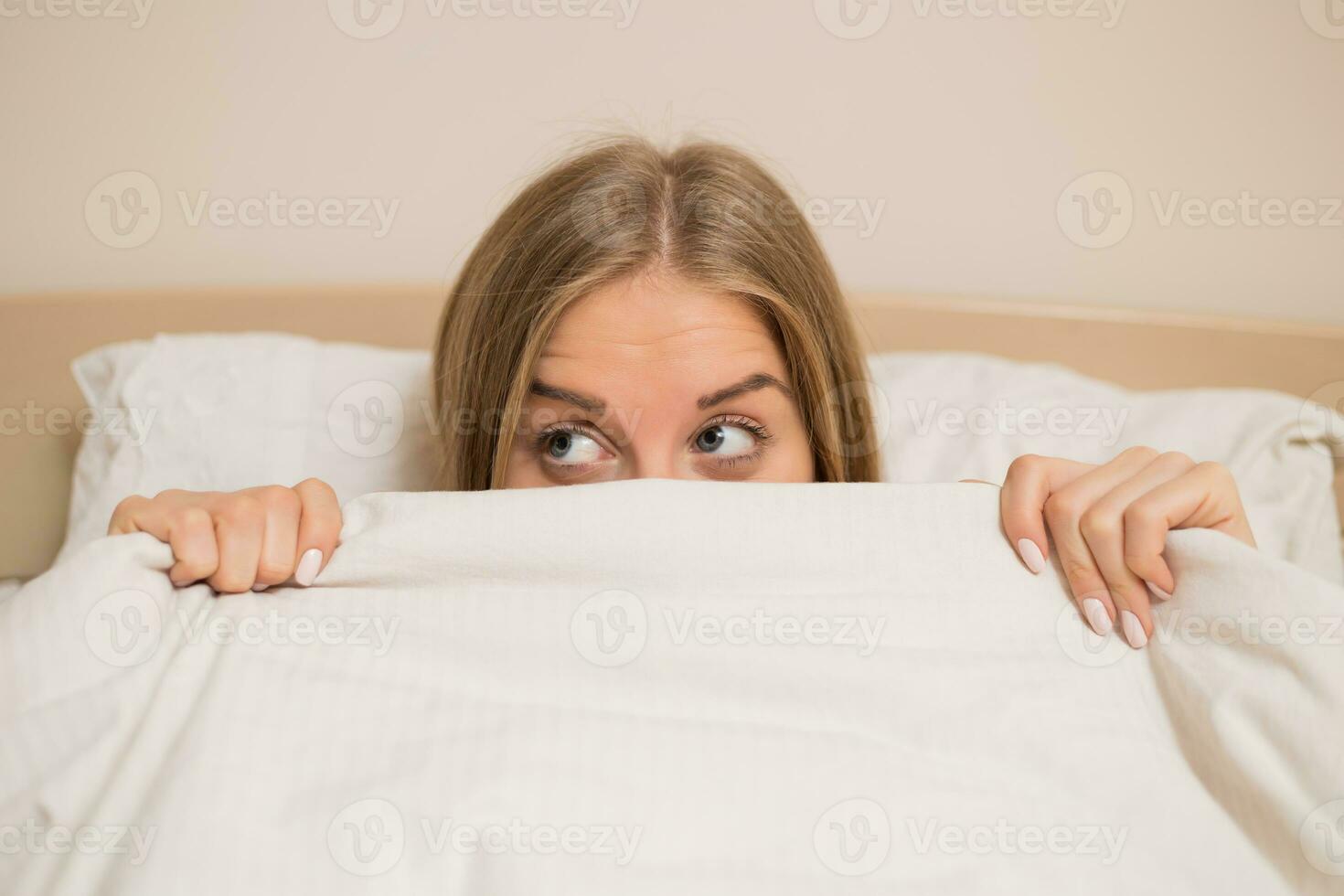 kvinna med skön blå ögon om i säng och kikar Bakom täcke foto