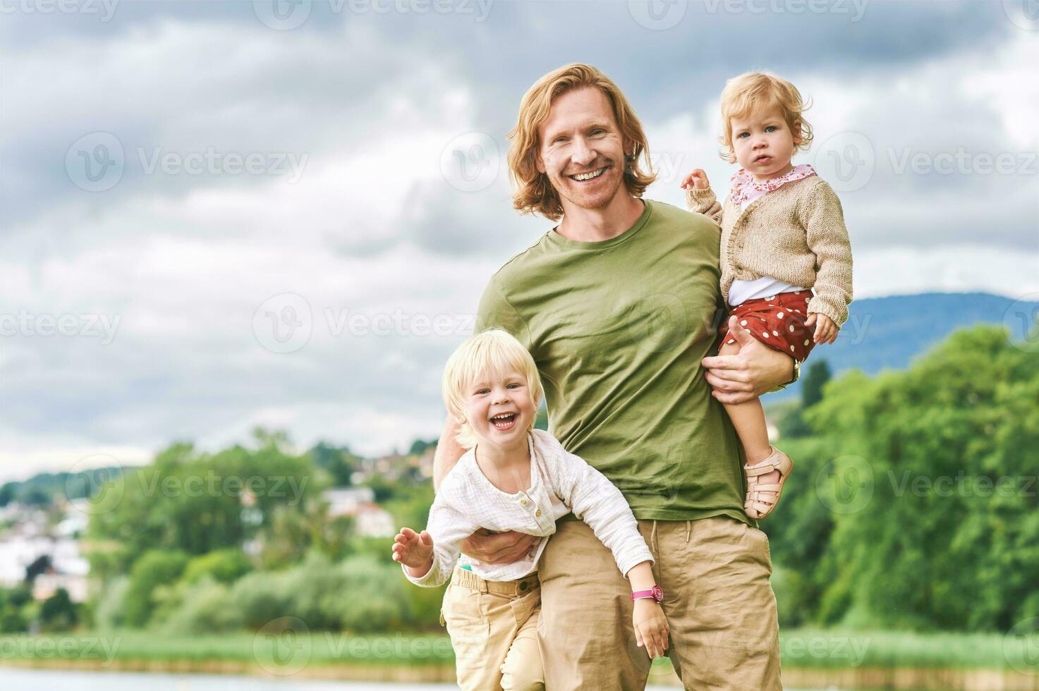 utomhus- porträtt av Lycklig ung far innehav två barn i vapen, litet barn flicka och preschooler pojke har roligt med pappa på en trevlig solig dag foto